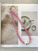Pink cat’s eye and baroque pearl necklace styled on an open magazine with neutral objects.