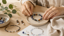 Elegant hands arranging natural gemstone jewelry with beads, earrings, sketch paper, linen, and ceramic tray in a calm mindful ritual setting.